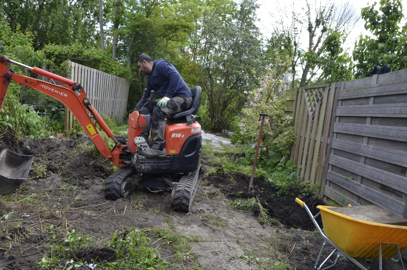 Hovenier graaft de tuin uit met de minigraafmachine, kruiwagen ernaast