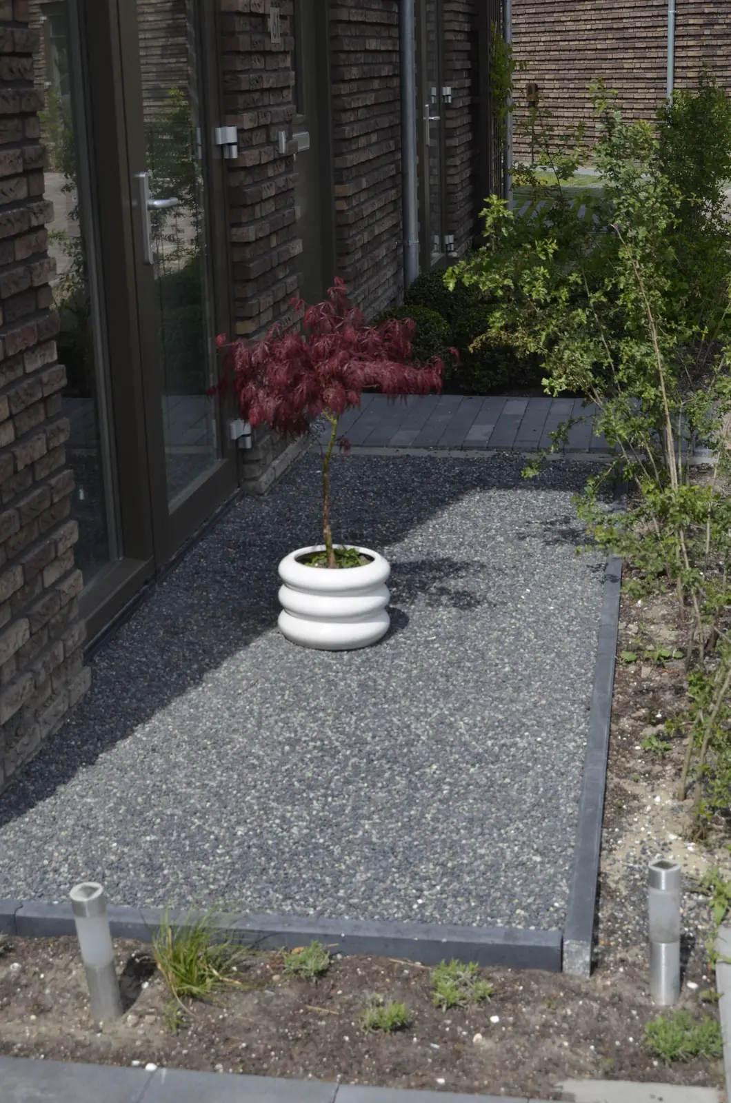 Zijtuin met grijs siergrind, een jonge esdoorn in pot en verlichting langs het pad