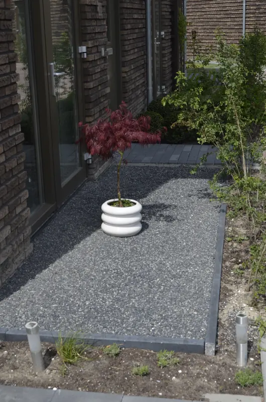 Zijtuin met grijs siergrind, een jonge esdoorn in pot en verlichting langs het pad